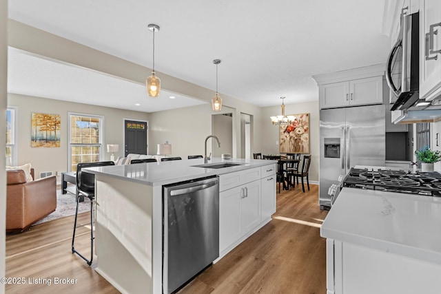 kitchen with appliances with stainless steel finishes, a kitchen island with sink, sink, white cabinetry, and hanging light fixtures