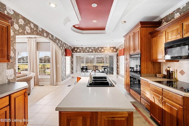 kitchen with a raised ceiling, a kitchen island with sink, black appliances, and ornamental molding