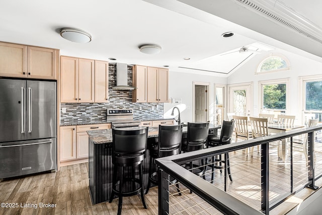 kitchen with appliances with stainless steel finishes, light brown cabinets, vaulted ceiling, and wall chimney range hood