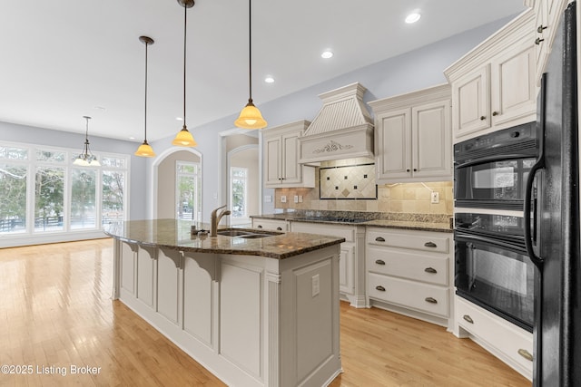 kitchen featuring backsplash, custom range hood, black appliances, dark stone countertops, and an island with sink