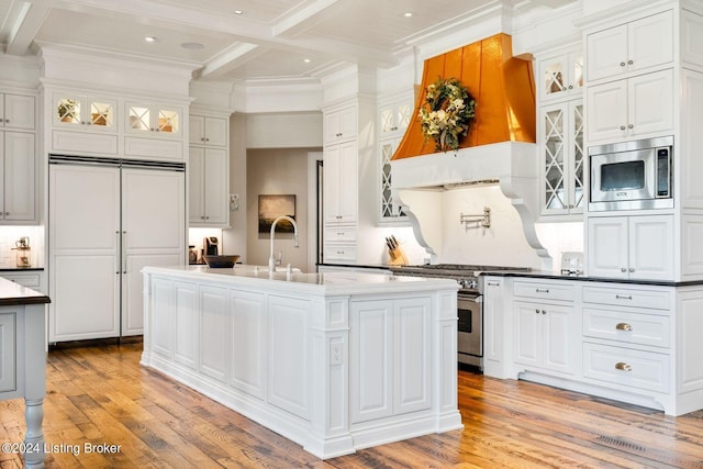 kitchen featuring coffered ceiling, beam ceiling, built in appliances, a center island with sink, and white cabinetry