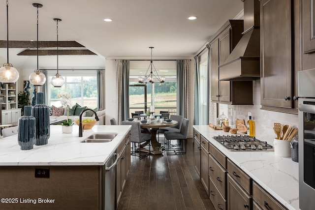 kitchen with pendant lighting, sink, an island with sink, appliances with stainless steel finishes, and custom range hood