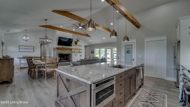 kitchen featuring a stone fireplace, an island with sink, ceiling fan, beamed ceiling, and sink