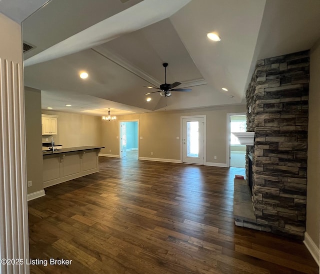 unfurnished living room featuring ceiling fan with notable chandelier, a raised ceiling, dark wood-type flooring, sink, and a stone fireplace