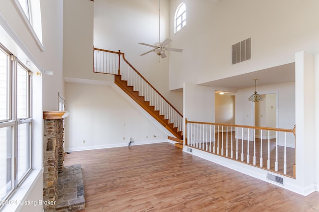 unfurnished living room featuring ceiling fan with notable chandelier, hardwood / wood-style floors, a towering ceiling, and a stone fireplace