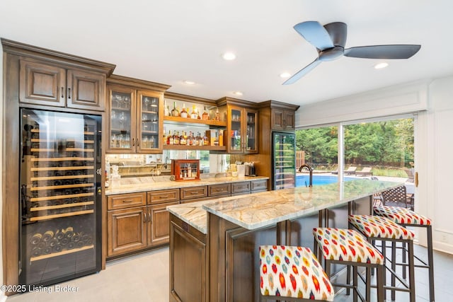 kitchen with an island with sink, wine cooler, light stone counters, glass insert cabinets, and a breakfast bar area