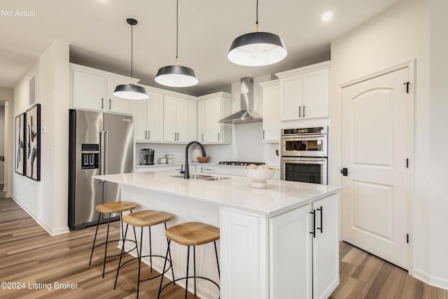 kitchen with appliances with stainless steel finishes, wall chimney exhaust hood, a kitchen island with sink, sink, and white cabinets