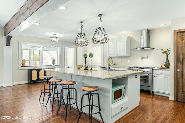 kitchen featuring light stone countertops, white cabinetry, wall chimney range hood, a center island with sink, and stainless steel range