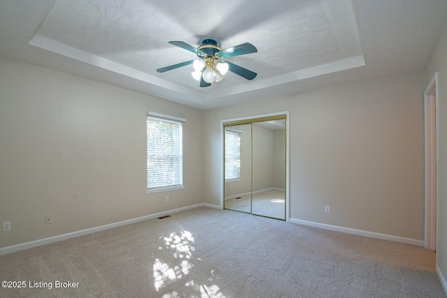 unfurnished bedroom featuring a textured ceiling, a closet, a raised ceiling, ceiling fan, and light colored carpet