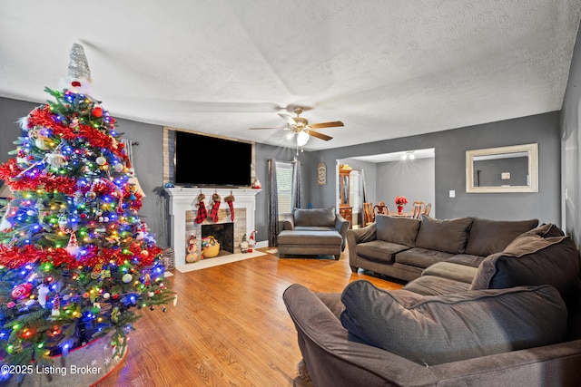 living room with a brick fireplace, a textured ceiling, ceiling fan, and light hardwood / wood-style flooring