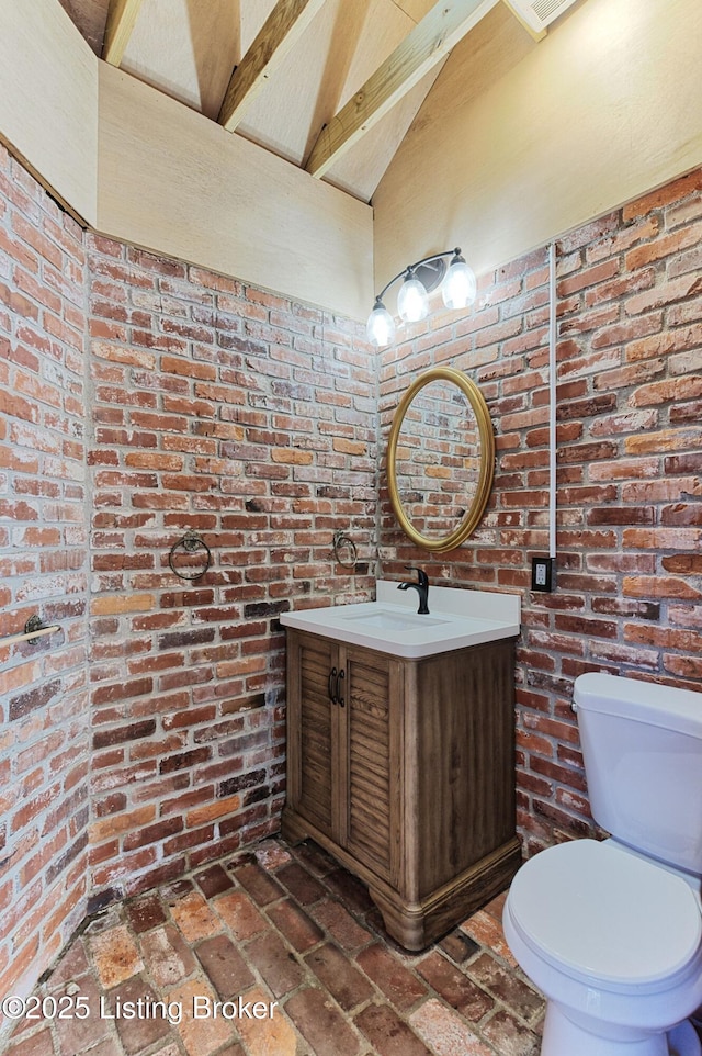 bathroom with vanity, vaulted ceiling with beams, toilet, and brick wall