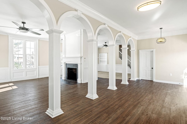 unfurnished living room featuring dark wood-type flooring, ornamental molding, ceiling fan with notable chandelier, and decorative columns