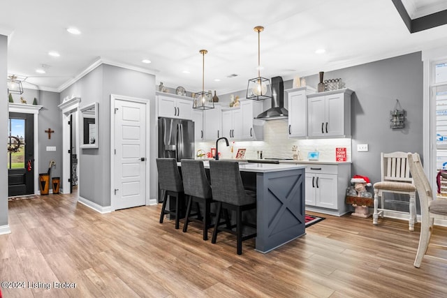 kitchen with a center island with sink, light hardwood / wood-style floors, hanging light fixtures, wall chimney range hood, and tasteful backsplash