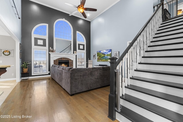 living room featuring a high ceiling, wood-type flooring, a fireplace, ceiling fan, and crown molding