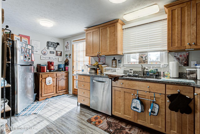 kitchen featuring appliances with stainless steel finishes, light wood-type flooring, dark stone countertops, a textured ceiling, and sink