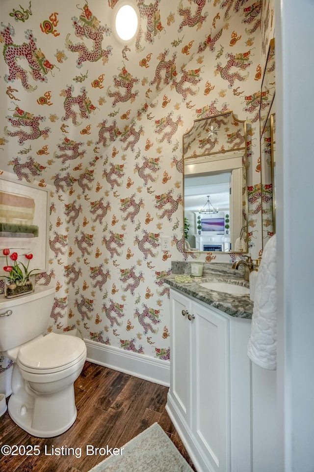 half bathroom with wallpapered walls, baseboards, toilet, wood finished floors, and vanity