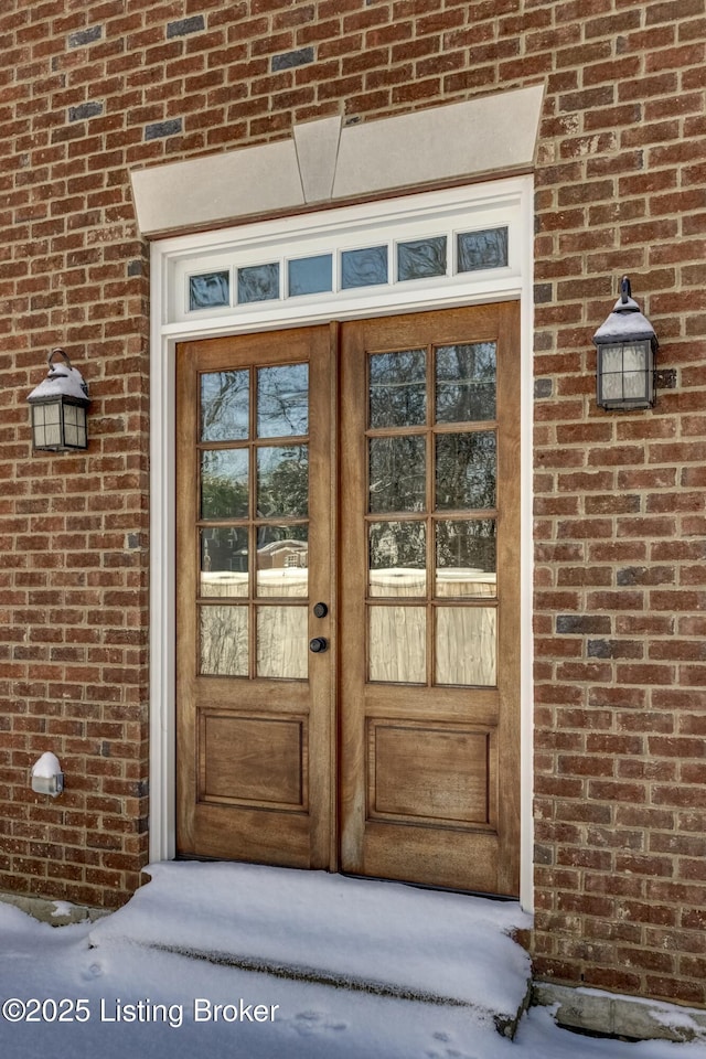 snow covered property entrance featuring french doors and brick siding