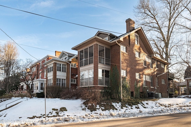 view of snow covered exterior with a sunroom, a chimney, cooling unit, and brick siding