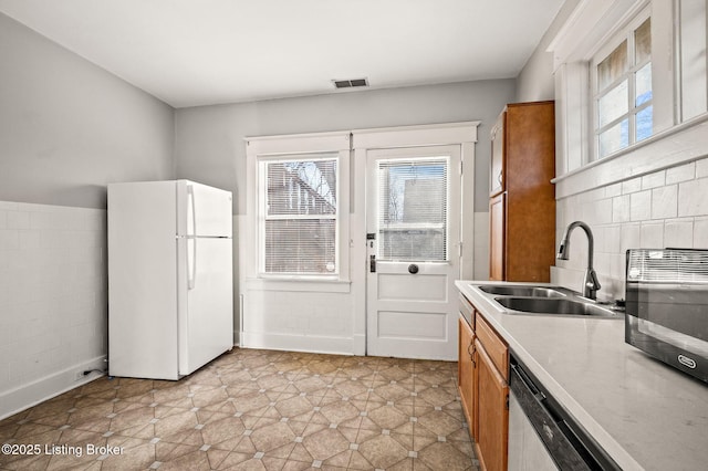 kitchen featuring visible vents, dishwashing machine, brown cabinets, freestanding refrigerator, and a sink