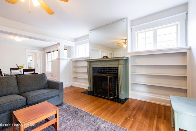 living area with a ceiling fan, a fireplace, built in shelves, and wood finished floors