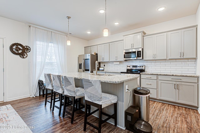 kitchen with pendant lighting, light stone counters, stainless steel appliances, and an island with sink