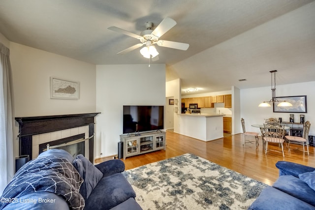 living room with ceiling fan, lofted ceiling, a tiled fireplace, and light wood-type flooring