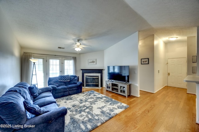 living room with a tiled fireplace, a textured ceiling, ceiling fan, and light hardwood / wood-style flooring