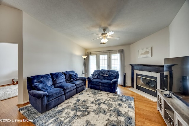 living room featuring ceiling fan, a tiled fireplace, a textured ceiling, and light wood-type flooring