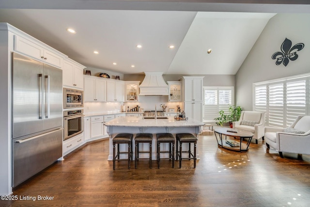 kitchen featuring custom range hood, tasteful backsplash, a center island with sink, a breakfast bar area, and built in appliances