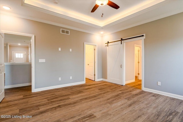 unfurnished bedroom with ornamental molding, a tray ceiling, hardwood / wood-style flooring, ceiling fan, and a barn door