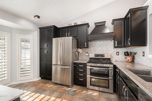 kitchen with vaulted ceiling, appliances with stainless steel finishes, tasteful backsplash, custom exhaust hood, and light stone countertops