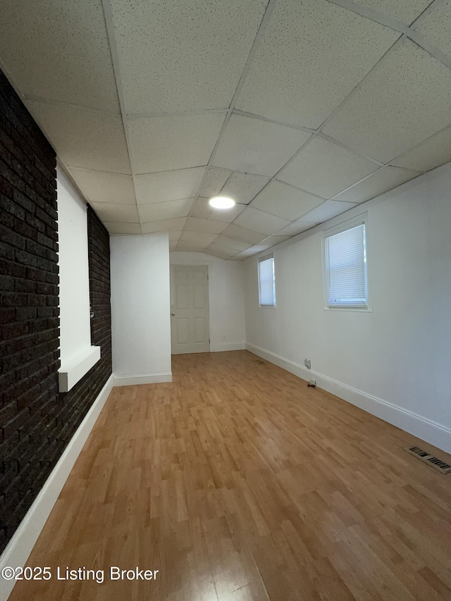 basement featuring a drop ceiling, light hardwood / wood-style flooring, and brick wall