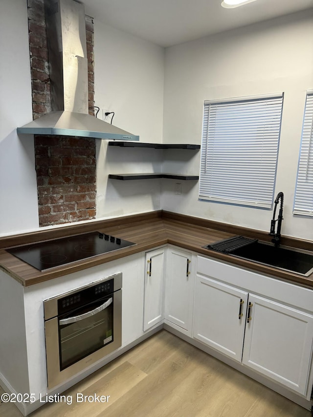 kitchen featuring black electric stovetop, open shelves, stainless steel oven, a sink, and wall chimney range hood