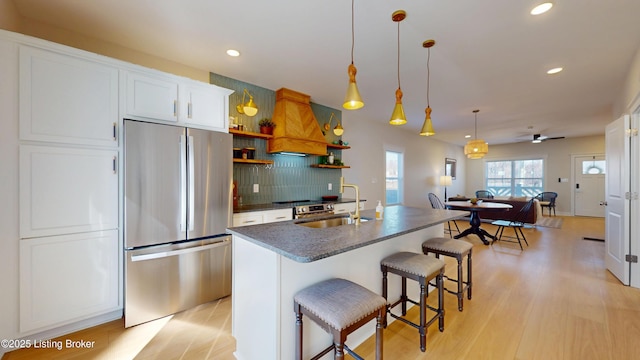 kitchen featuring dark countertops, an island with sink, a breakfast bar area, stainless steel refrigerator, and white cabinetry