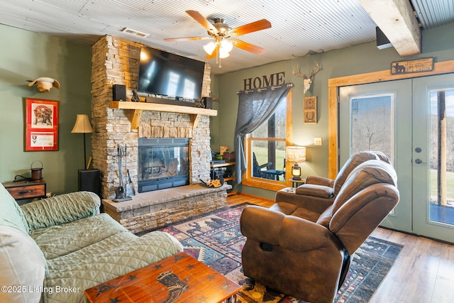 living room with french doors, a stone fireplace, beamed ceiling, ceiling fan, and light hardwood / wood-style floors