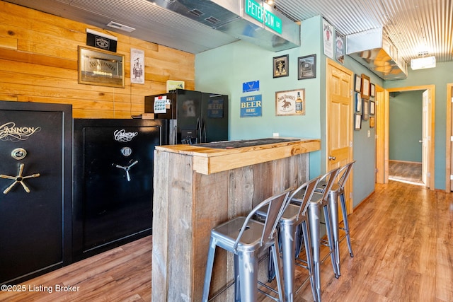 bar with wood counters, black refrigerator with ice dispenser, light hardwood / wood-style floors, and wood walls