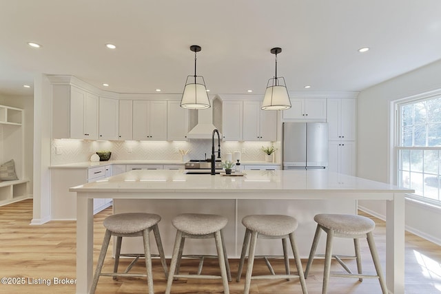 kitchen featuring white cabinetry, decorative light fixtures, stainless steel refrigerator, an island with sink, and light hardwood / wood-style floors