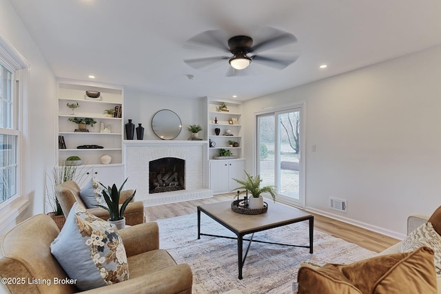 living room with a brick fireplace, built in features, light hardwood / wood-style floors, and ceiling fan