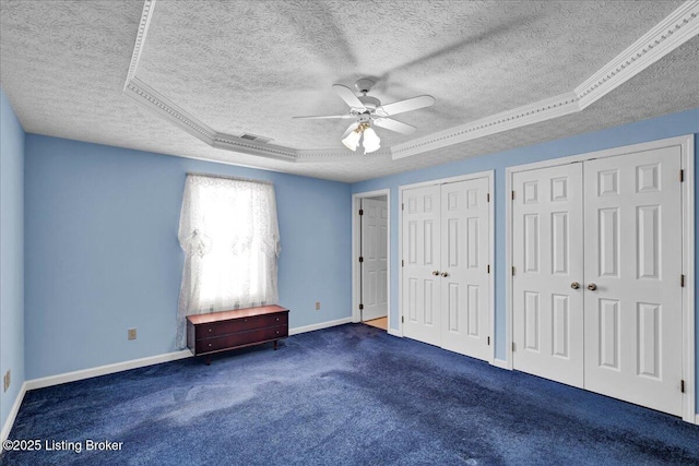 unfurnished bedroom featuring ceiling fan, dark colored carpet, a tray ceiling, a textured ceiling, and two closets