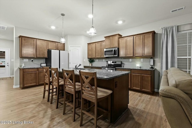 kitchen featuring a kitchen bar, decorative light fixtures, appliances with stainless steel finishes, dark stone counters, and a kitchen island with sink