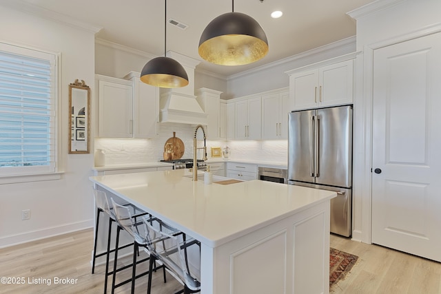 kitchen with crown molding, stainless steel refrigerator, pendant lighting, a kitchen island with sink, and white cabinets