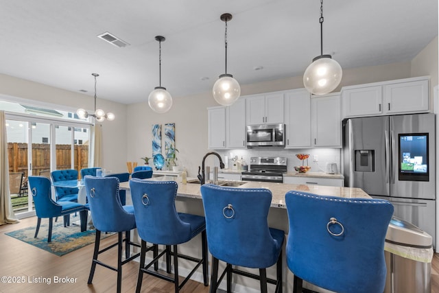 kitchen with hanging light fixtures, appliances with stainless steel finishes, an island with sink, light stone countertops, and white cabinets