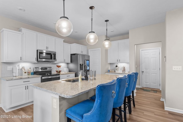 kitchen featuring pendant lighting, stainless steel appliances, light stone countertops, white cabinets, and a center island with sink