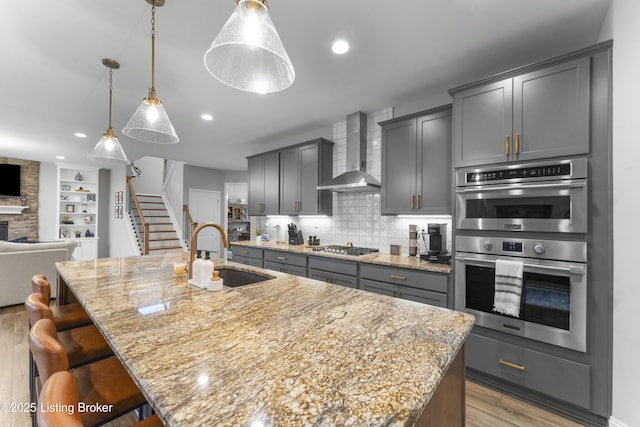 kitchen featuring appliances with stainless steel finishes, sink, hanging light fixtures, a large island with sink, and wall chimney range hood