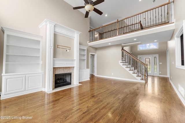 unfurnished living room with built in shelves, wood-type flooring, ceiling fan, a towering ceiling, and a fireplace