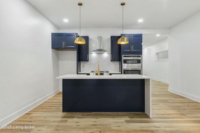 kitchen featuring blue cabinets, an island with sink, tasteful backsplash, light wood-type flooring, and wall chimney exhaust hood