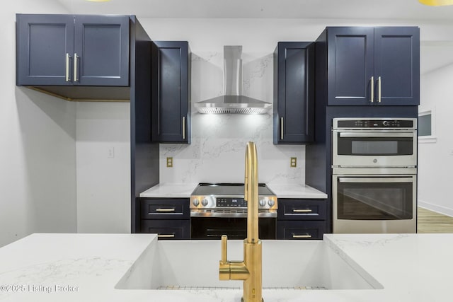 kitchen featuring wall chimney exhaust hood, blue cabinets, light stone counters, appliances with stainless steel finishes, and backsplash