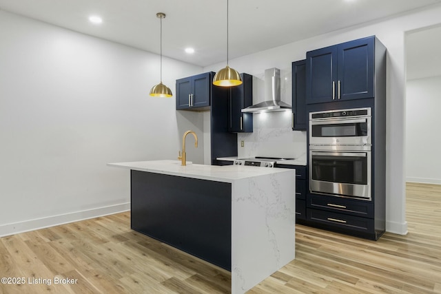 kitchen featuring sink, stainless steel double oven, a kitchen island with sink, light stone counters, and wall chimney range hood