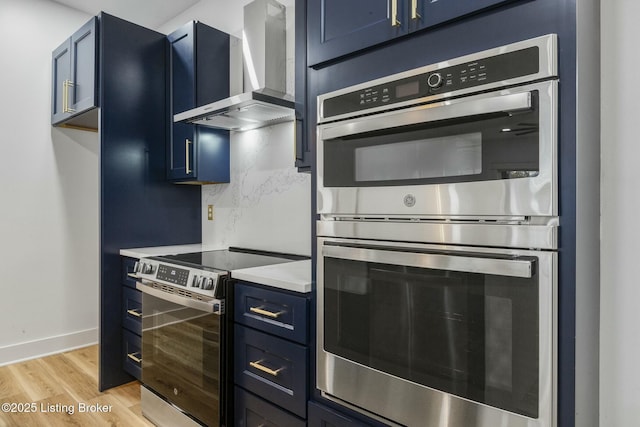 kitchen featuring tasteful backsplash, blue cabinets, light hardwood / wood-style floors, stainless steel appliances, and wall chimney range hood