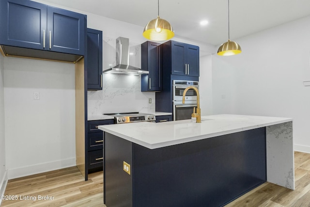 kitchen with wall chimney exhaust hood, blue cabinets, hanging light fixtures, appliances with stainless steel finishes, and an island with sink